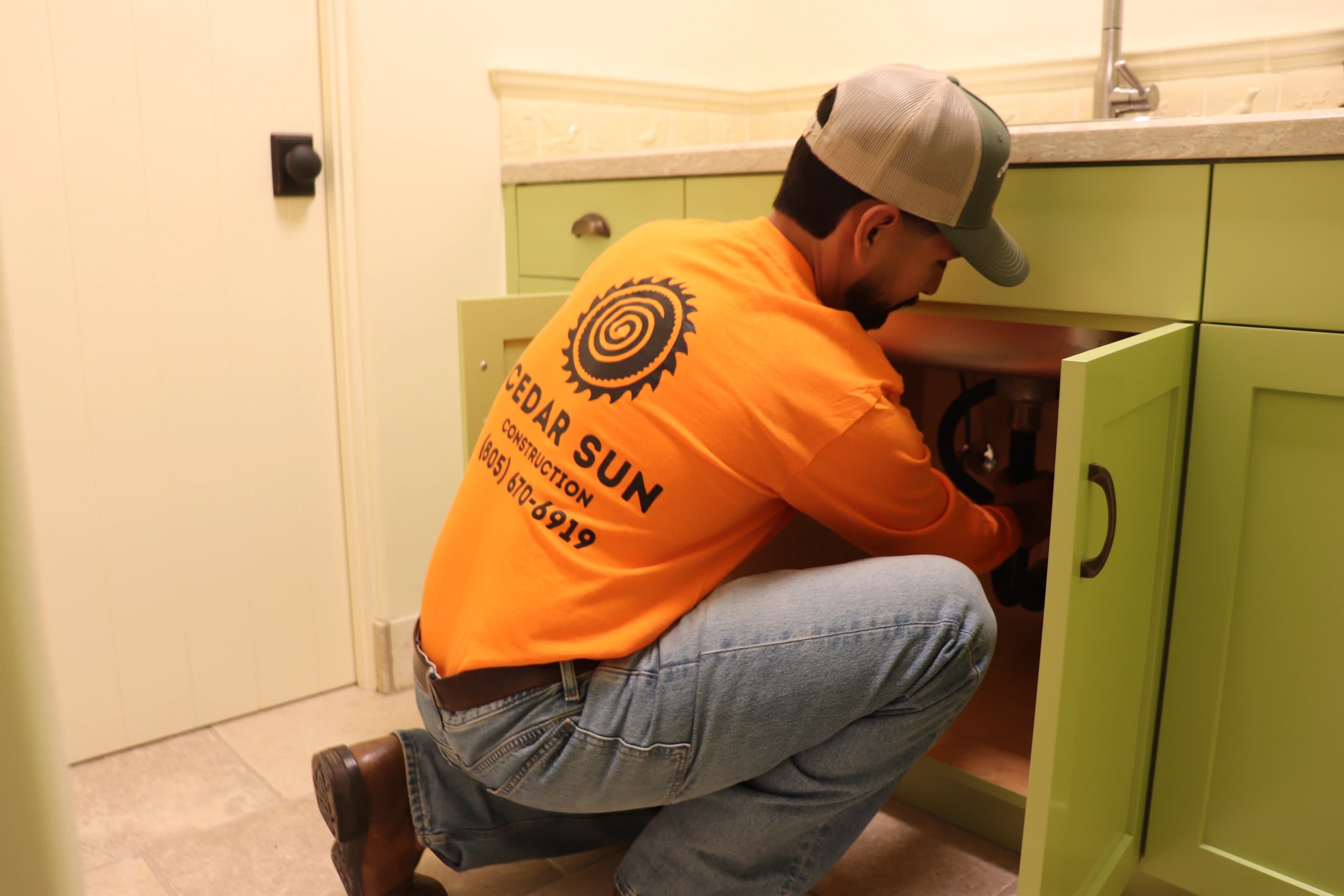 Cedar Sun Construction owner in a Cedar Sun Construction work tee installing plumbing under a sage green vanity in Ventura County, CA